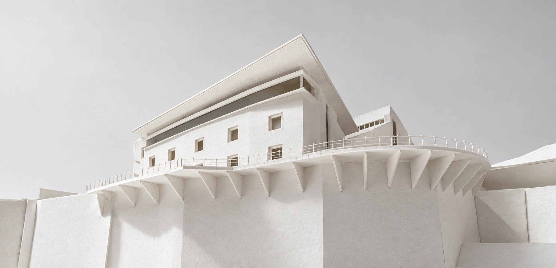 Visualización editorial de una maqueta arquitectónica de cartón blanco con cubierta inclinada, plataforma curva y soportes triangulares